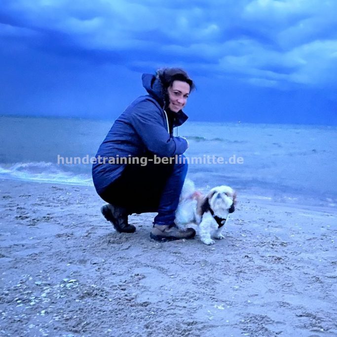 Katja mit Hündin Cindy am Ostseestrand. Cindy ist eine Shi-Tzu Hündind. Katja kniet neben ihr und schaut in die Kamera.